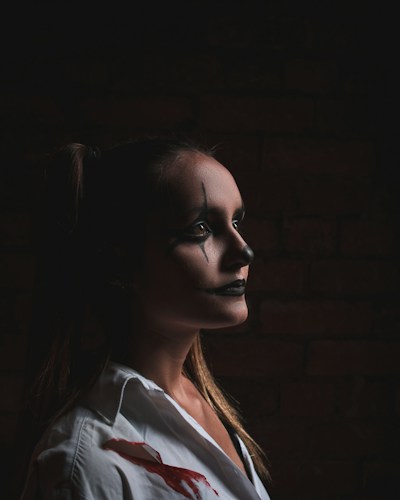 A person with dark, theatrical makeup is captured in profile against a dimly lit background. The makeup features exaggerated black lines and a stark white base, giving a dramatic, intense look. The individual is wearing a white garment, possibly a shirt, with noticeable red stains, enhancing the dramatic and possibly eerie atmosphere.