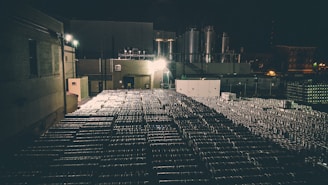A dimly lit industrial area with numerous stacked metal barrels extending across the scene. Large industrial tanks and a structure with brick walls are visible in the background, illuminated by artificial lights, creating a shadowy and moody atmosphere.