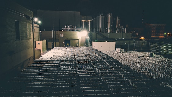 A dimly lit industrial area with numerous stacked metal barrels extending across the scene. Large industrial tanks and a structure with brick walls are visible in the background, illuminated by artificial lights, creating a shadowy and moody atmosphere.