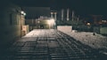 A dimly lit industrial area with numerous stacked metal barrels extending across the scene. Large industrial tanks and a structure with brick walls are visible in the background, illuminated by artificial lights, creating a shadowy and moody atmosphere.