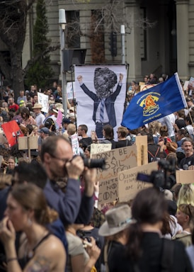 A large crowd of people is participating in a protest. Many are holding signs with various slogans related to climate change, such as 'Don't be a fossil fool' and 'Oceans are rising'. A prominent sign depicts a person in a suit with a rock for a head. The crowd is diverse, with some individuals carrying cameras and others wearing hats. There is also a blue flag prominently displayed in the center.