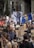 A large crowd of people is participating in a protest. Many are holding signs with various slogans related to climate change, such as 'Don't be a fossil fool' and 'Oceans are rising'. A prominent sign depicts a person in a suit with a rock for a head. The crowd is diverse, with some individuals carrying cameras and others wearing hats. There is also a blue flag prominently displayed in the center.