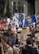 A large crowd of people is participating in a protest. Many are holding signs with various slogans related to climate change, such as 'Don't be a fossil fool' and 'Oceans are rising'. A prominent sign depicts a person in a suit with a rock for a head. The crowd is diverse, with some individuals carrying cameras and others wearing hats. There is also a blue flag prominently displayed in the center.
