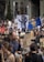 A large crowd of people is participating in a protest. Many are holding signs with various slogans related to climate change, such as 'Don't be a fossil fool' and 'Oceans are rising'. A prominent sign depicts a person in a suit with a rock for a head. The crowd is diverse, with some individuals carrying cameras and others wearing hats. There is also a blue flag prominently displayed in the center.