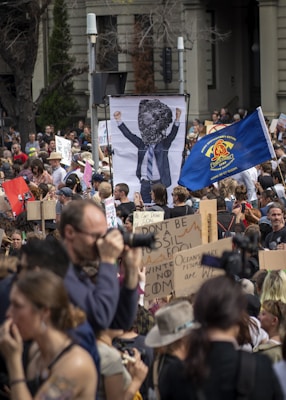 A large crowd of people is participating in a protest. Many are holding signs with various slogans related to climate change, such as 'Don't be a fossil fool' and 'Oceans are rising'. A prominent sign depicts a person in a suit with a rock for a head. The crowd is diverse, with some individuals carrying cameras and others wearing hats. There is also a blue flag prominently displayed in the center.