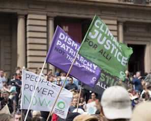 A large group of people gather outside a grand building with columns, holding various protest signs and banners. Green and purple flags display messages about climate justice, dismantling patriarchy, and empowering people.