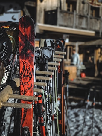 A collection of skis and ski poles are neatly arranged in a wooden rack. The skis feature various designs and colors, with one prominently displaying a vivid red and black pattern. The background shows a rustic wooden building, possibly a chalet, with snow visible on the ground.