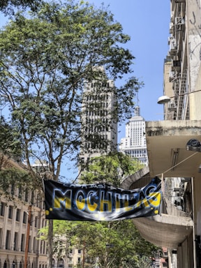 A street scene with tall buildings partially obscured by lush green trees. A prominent banner displaying the word 'MOCHILAS' in vibrant yellow and blue hangs across the street. Sunlight casts bright highlights, creating a contrast against the shadows of the buildings and trees.