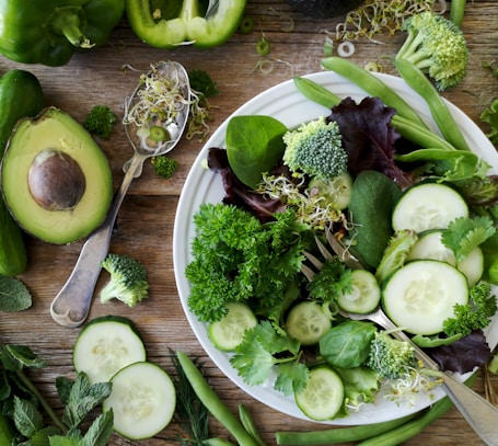 sliced broccoli and cucumber on plate with gray stainless steel fork near green bell pepper, snowpea, and avocado fruit