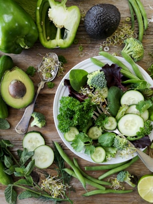sliced broccoli and cucumber on plate with gray stainless steel fork near green bell pepper, snowpea, and avocado fruit