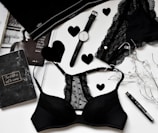 A flat lay of black lingerie and accessories on a white surface, including a watch, a journal with 'The best is yet to come' written on it, a magazine partially visible, and decorative elements like black hearts and string lights.
