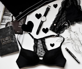 A flat lay of black lingerie and accessories on a white surface, including a watch, a journal with 'The best is yet to come' written on it, a magazine partially visible, and decorative elements like black hearts and string lights.