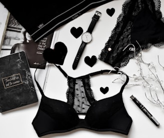 A flat lay of black lingerie and accessories on a white surface, including a watch, a journal with 'The best is yet to come' written on it, a magazine partially visible, and decorative elements like black hearts and string lights.