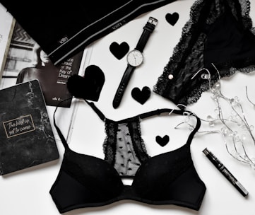 A flat lay of black lingerie and accessories on a white surface, including a watch, a journal with 'The best is yet to come' written on it, a magazine partially visible, and decorative elements like black hearts and string lights.
