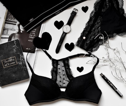 A flat lay of black lingerie and accessories on a white surface, including a watch, a journal with 'The best is yet to come' written on it, a magazine partially visible, and decorative elements like black hearts and string lights.