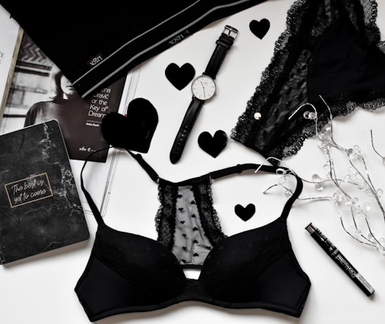 A flat lay of black lingerie and accessories on a white surface, including a watch, a journal with 'The best is yet to come' written on it, a magazine partially visible, and decorative elements like black hearts and string lights.