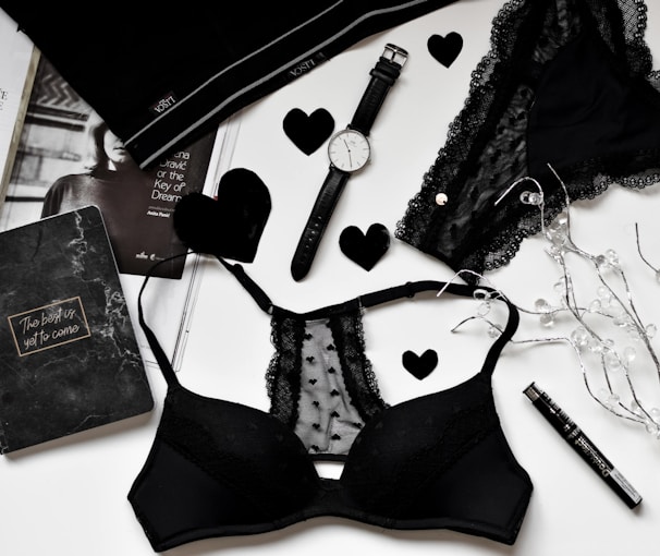 A flat lay of black lingerie and accessories on a white surface, including a watch, a journal with 'The best is yet to come' written on it, a magazine partially visible, and decorative elements like black hearts and string lights.