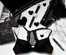 A flat lay of black lingerie and accessories on a white surface, including a watch, a journal with 'The best is yet to come' written on it, a magazine partially visible, and decorative elements like black hearts and string lights.