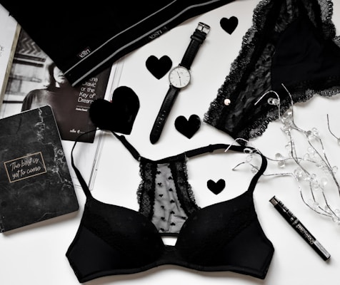 A flat lay of black lingerie and accessories on a white surface, including a watch, a journal with 'The best is yet to come' written on it, a magazine partially visible, and decorative elements like black hearts and string lights.