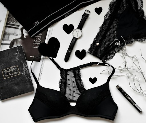 A flat lay of black lingerie and accessories on a white surface, including a watch, a journal with 'The best is yet to come' written on it, a magazine partially visible, and decorative elements like black hearts and string lights.