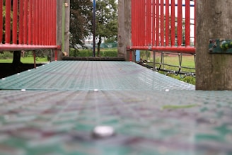 A playground bridge with metal grating and red railings extends forward, surrounded by greenery in the background. The bridge appears to be part of a larger play structure.