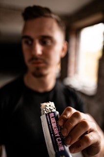 A person holds a protein bar in their hand, with a label reading 'Blueberry Cake'. The focus is on the hand and the bar, while the person&rsquo;s face is slightly out of focus in the background. The setting appears to be indoors with a window in the background allowing natural light.