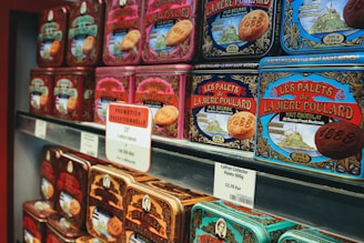 Rows of colorful vintage-style tin boxes are displayed on shelves, each containing La Mere Poulard butter cookies. The packaging features intricate designs and illustrations, emphasizing a traditional and artisanal product. Prominent signage with pricing and promotional offers is visible, indicating a special sale.