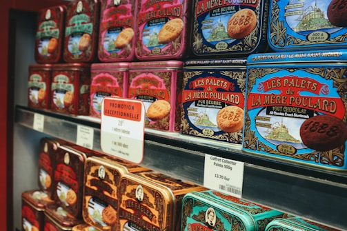 Rows of colorful vintage-style tin boxes are displayed on shelves, each containing La Mere Poulard butter cookies. The packaging features intricate designs and illustrations, emphasizing a traditional and artisanal product. Prominent signage with pricing and promotional offers is visible, indicating a special sale.