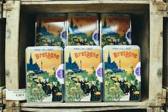A stack of tin boxes with vintage-style artwork featuring the region of Bretagne, showing a village scene with a cathedral, people in traditional clothing, and yellow flowers. The boxes are displayed on wooden shelves with a price tag indicating the product, which is chocolate.