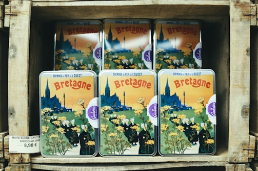 A stack of tin boxes with vintage-style artwork featuring the region of Bretagne, showing a village scene with a cathedral, people in traditional clothing, and yellow flowers. The boxes are displayed on wooden shelves with a price tag indicating the product, which is chocolate.