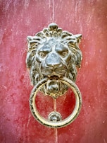 A metal lion's head door knocker with intricate detailing, mounted on a red, weathered wooden door. The lion's mouth holds a large, circular metal ring.