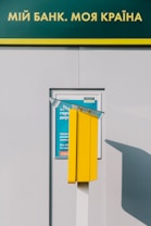 A yellow kiosk-like structure stands in front of a bank advertisement. The advertisement is framed and blue with text partially visible through a reflective glass. Above, there is a green and yellow sign with white text in Cyrillic.