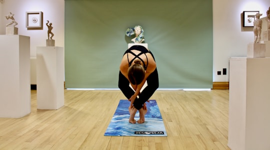 A person is practicing yoga on a blue mat in an art gallery setting. The room has wooden floors and is decorated with sculptures placed on pedestals. The background features a green partition and a bust sculpture. The lighting is soft, creating a serene ambiance.