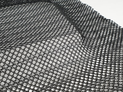 A detailed black mesh material with intricate patterns is laid out, displaying a grid of interlocking loops and open spaces. The texture appears flexible and lightweight, with subtle shadows adding depth.