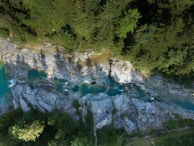 An aerial view of a lush green forest surrounding a rocky riverbed with clear turquoise pools of water. The landscape features rich foliage and a mix of large, smooth boulders along the river.