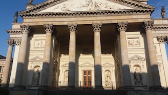 A classical building facade featuring tall columns with ornate capitals. Statues are placed in niches on the pediment and above the entrance. The architecture is detailed with reliefs and decorative elements, lending a sense of grandeur. The building appears to be basked in warm sunlight, casting shadows that enhance the depth and textures of the structure.