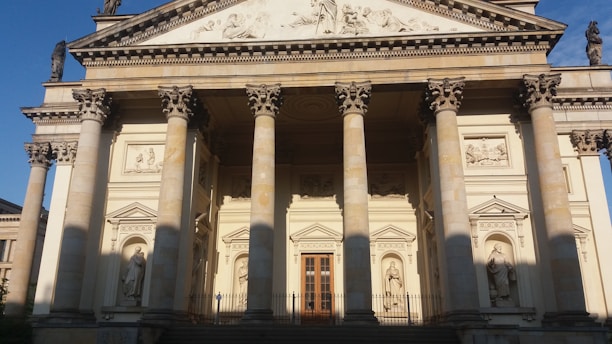 A classical building facade featuring tall columns with ornate capitals. Statues are placed in niches on the pediment and above the entrance. The architecture is detailed with reliefs and decorative elements, lending a sense of grandeur. The building appears to be basked in warm sunlight, casting shadows that enhance the depth and textures of the structure.