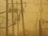 A network of electric power lines and poles is set against a hazy, yellow-tinted sky. The silhouettes of the electrical equipment create a complex web of lines and shapes. Vegetation is visible at the bottom, and there is a metal fence on the right side of the image.