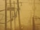A network of electric power lines and poles is set against a hazy, yellow-tinted sky. The silhouettes of the electrical equipment create a complex web of lines and shapes. Vegetation is visible at the bottom, and there is a metal fence on the right side of the image.
