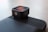 A sleek, black square-shaped camera lens accessory with a glass top displaying subtle reflections. It is mounted on a textured black base and situated on a smooth, neutral-colored surface.