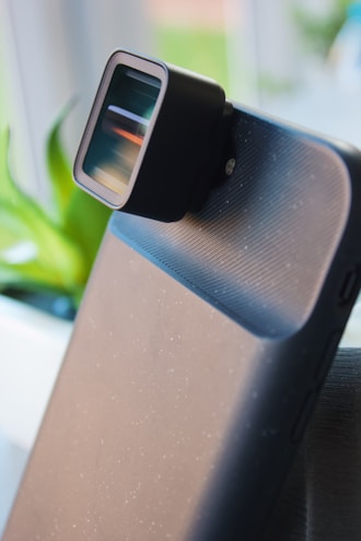A close-up view of a smartphone fitted with an external camera lens. The lens is rectangular and attached to the phone's camera area. There is a soft-focus background with a plant visible.
