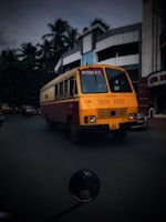 A brightly colored bus with a distinctive yellow and red design is driving down a street lined with modern buildings. Palm trees can be seen in the background, and there are a few other vehicles on the road. The bus has the letters 'KSRTC' on the front and a license plate with the number 'KLX 6265'.