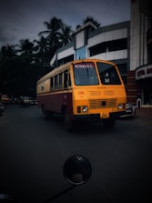 A brightly colored bus with a distinctive yellow and red design is driving down a street lined with modern buildings. Palm trees can be seen in the background, and there are a few other vehicles on the road. The bus has the letters 'KSRTC' on the front and a license plate with the number 'KLX 6265'.