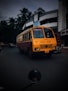 A brightly colored bus with a distinctive yellow and red design is driving down a street lined with modern buildings. Palm trees can be seen in the background, and there are a few other vehicles on the road. The bus has the letters 'KSRTC' on the front and a license plate with the number 'KLX 6265'.