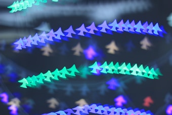 A visually striking pattern of bokeh lights resembling small airplane silhouettes, arranged in a cascading diagonal formation. The colors shift between shades of blue, green, and aqua against a dark background, creating a soft, dreamy aesthetic.