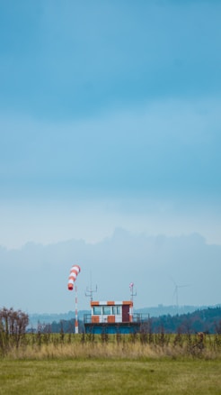 A small airfield control building with a striped windsock stands amidst green grass under a vast blue sky. A distant wind turbine is faintly visible on the horizon surrounded by trees and bushes.