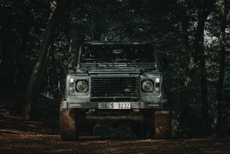 A rugged off-road vehicle is parked in a dense forest setting, surrounded by towering trees and shaded areas. The vehicle's headlights are on, and it is covered with dirt and mud, indicating it has been driven through rough terrain.