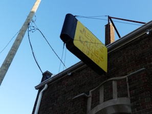 A yellow and black sign with raised letters saying 'JESUS CARES DO YOU' is mounted on the side of a brick building. The sign is supported by metal rods and is placed above an archway with decorative brickwork. Overhead wires are visible, connecting to a utility pole. The sky is a clear blue, suggesting a sunny day.
