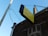 A yellow and black sign with raised letters saying 'JESUS CARES DO YOU' is mounted on the side of a brick building. The sign is supported by metal rods and is placed above an archway with decorative brickwork. Overhead wires are visible, connecting to a utility pole. The sky is a clear blue, suggesting a sunny day.