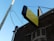 A yellow and black sign with raised letters saying 'JESUS CARES DO YOU' is mounted on the side of a brick building. The sign is supported by metal rods and is placed above an archway with decorative brickwork. Overhead wires are visible, connecting to a utility pole. The sky is a clear blue, suggesting a sunny day.